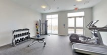 This fitness center is a thoughtfully designed workout space that provides a comprehensive range of exercise options within a clean, modern environment. The room features durable rubber flooring throughout, offering both safety and functionality for various types of exercise. Large floor-to-ceiling windows and a set of double glass doors flood the space with natural light while offering views of an outdoor terrace area, creating an open and motivating atmosphere.

The cardio section is well-appointed, featuring multiple treadmills and an elliptical machine, each paired with wall-mounted flat-screen televisions for entertainment during workouts. Complementing the cardio equipment is a rowing machine, providing users with a full range of aerobic options. For strength training, the space includes a multi-function cable machine, a dedicated free weights area with a tiered dumbbell rack offering a wide selection of weights, a medicine ball rack with assorted sizes, and an adjustable workout bench.

The neutral white walls, flush-mounted ceiling lights, and clearly marked exit signage contribute to the room's clean, functional aesthetic. A mirrored wall section enhances the sense of space and supports form-checking during exercise. Access points on both ends of the room ensure convenient entry and exit for users.