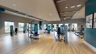 This fitness center at Sonceto Apartments is a thoughtfully designed, single-floor facility that offers a comprehensive workout experience. The open floor plan is finished with light hardwood-style flooring and accented by bold teal feature walls that provide a vibrant, energizing atmosphere throughout the space.

The cardio zone is lined with multiple treadmills and an elliptical trainer, each equipped with individual display screens, all positioned in front of a row of large windows that frame lush outdoor views. Nearby, a dedicated strength training area houses a full suite of Matrix-brand cable and resistance machines, providing options for a wide range of muscle groups.

A functional training corner features battle ropes, a full dumbbell rack with varying weights, and medicine balls, supporting dynamic and free-weight workouts. A wall-mounted flat-screen television keeps members entertained or informed during training sessions. An additional area includes a Kinomap smart board and step platforms, ideal for interactive and low-impact training.

Recessed lighting and ceiling fans ensure consistent illumination and airflow, while multiple windows on all sides flood the space with natural light and views of the surrounding grounds. Entry is through sleek black-framed glass double doors, creating a polished and welcoming first impression. The overall ambiance is modern, clean, and purpose-built for an active lifestyle.