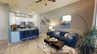 This single-floor space showcases a thoughtfully designed open-concept layout that seamlessly connects the kitchen and living areas, creating a cohesive and functional living environment. The kitchen is outfitted with stainless steel appliances, including a French door refrigerator and range, and features a bold navy blue island with granite countertops and seating for multiple bar-height chairs — an ideal spot for casual dining or entertaining. Pendant lighting hangs above the island, complementing the modern white cabinetry and stylish tile backsplash. The living area exudes a contemporary yet comfortable ambiance, anchored by light wood-look flooring that runs throughout the space. Vaulted ceilings with a ceiling fan add a sense of openness and airiness, while recessed lighting ensures the space is well-lit. Neutral gray walls serve as a clean backdrop for curated décor, including an arrangement of circular wall mirrors and a vibrant cityscape painting. The space benefits from an efficient, flowing layout that balances style and livability, making it equally suited for relaxing and hosting guests.