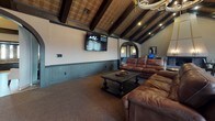 Located at Fountain Pointe in Grand Blanc, Michigan, this single-floor clubhouse offers a well-appointed gathering space defined by distinctive architectural character and functional amenities throughout.

The centerpiece of the space is a dramatic great room with soaring vaulted ceilings accented by exposed dark wood beams and a decorative tin ceiling inlay. A grand brick fireplace with a bold angular surround commands one wall, serving as a striking focal point. The room accommodates a generous lounge area with comfortable leather sectional seating and a wall-mounted flat-screen television, ideal for group gatherings and presentations.

Adjacent to the lounge, a dining area features a round table with leather chairs beneath the same impressive beamed ceiling. Large sliding glass doors flood this area with natural light and provide direct access to an outdoor deck, offering views of the surrounding landscape.

A workstation area with a desktop computer is conveniently integrated into the space, supporting resident services and administrative needs. A decorative dark wood built-in shelving unit serves as an elegant room divider, adding architectural interest while defining the transition between spaces.

The full kitchen is finished with warm cabinetry, granite countertops, light wood-style vinyl flooring, stainless steel appliances, and a pass-through bar counter — well-equipped to support events and everyday resident use.