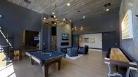 This sophisticated single-floor amenity lounge is designed with a bold, upscale industrial aesthetic that blends comfort and entertainment seamlessly. The space is anchored by a full-size billiards table finished in deep navy felt, positioned beneath two striking branch-style chandelier fixtures with exposed Edison bulbs that serve as dramatic focal points. The ceiling features a distinctive wood plank treatment that adds warmth and texture, complementing the polished concrete-style walls in a rich charcoal tone.

A wall-mounted flat-screen television is centered above a sleek linear fireplace, flanked by a floor-to-ceiling stacked firewood display that adds a rustic, organic element to the otherwise modern space. Adjacent to the fireplace, a curated seating arrangement with plush dark upholstered chairs is set atop a geometric patterned area rug, creating an intimate conversation area.

Large-format tile flooring in a warm neutral tone spans the entire space, contributing to the open, airy feel accentuated by soaring ceiling heights. Recessed lighting supplements the pendant fixtures, ensuring the space is well-lit throughout. Artwork adorns the walls, adding a gallery-like quality. Additional gaming or activity areas are visible in the background, reinforcing this lounge's role as a comprehensive social and recreational destination.