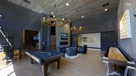 This sophisticated single-floor amenity lounge is designed with a bold, upscale industrial aesthetic that blends comfort and entertainment seamlessly. The space is anchored by a full-size billiards table finished in deep navy felt, positioned beneath two striking branch-style chandelier fixtures with exposed Edison bulbs that serve as dramatic focal points. The ceiling features a distinctive wood plank treatment that adds warmth and texture, complementing the polished concrete-style walls in a rich charcoal tone.

A wall-mounted flat-screen television is centered above a sleek linear fireplace, flanked by a floor-to-ceiling stacked firewood display that adds a rustic, organic element to the otherwise modern space. Adjacent to the fireplace, a curated seating arrangement with plush dark upholstered chairs is set atop a geometric patterned area rug, creating an intimate conversation area.

Large-format tile flooring in a warm neutral tone spans the entire space, contributing to the open, airy feel accentuated by soaring ceiling heights. Recessed lighting supplements the pendant fixtures, ensuring the space is well-lit throughout. Artwork adorns the walls, adding a gallery-like quality. Additional gaming or activity areas are visible in the background, reinforcing this lounge's role as a comprehensive social and recreational destination.