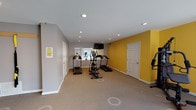 The Willowbrook fitness center is a thoughtfully designed, single-floor exercise facility that provides residents with a comprehensive range of workout options. The space features a bright, open layout accented by bold yellow and neutral gray walls, creating an energizing atmosphere conducive to physical activity. Recessed lighting throughout ensures the space is well-lit, while large glass doors along one wall flood the interior with natural light and provide views of the surrounding outdoor grounds.

The cardio zone is equipped with multiple treadmills and an elliptical machine, offering ample options for aerobic exercise. A multi-station strength training machine and a rowing machine provide additional full-body workout capabilities, while TRX suspension straps mounted to the wall cater to bodyweight and functional fitness training. A wall-mounted flat-screen television adds entertainment value during workouts.

A built-in water fountain is conveniently located near the exit for easy hydration. The carpeted flooring provides comfort underfoot, and clearly marked floor indicators help users maintain proper spacing between equipment. Additional doors within the facility provide access to utility or storage areas. The overall layout is spacious and well-organized, making the Willowbrook fitness center a practical and inviting amenity for daily exercise.