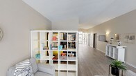 This single-floor space serves as a welcoming common area designed for relaxation and casual interaction. The layout is open and functional, with light-toned walls and smooth laminate wood flooring that runs throughout, contributing to a clean, contemporary aesthetic.

A central feature of the space is a large, white open-shelf unit that acts as a room divider, stocked with board games, books, and various items for resident use — including recognizable titles such as Yahtzee. Adjacent to this shelving area, vending machines provide convenient access to snacks and beverages. A comfortable seating area with upholstered furniture is positioned nearby, creating an informal lounge setting.

The entry zone features floor-to-ceiling glazed panels and a glass door that flood the area with natural light while offering views of the surrounding suburban neighborhood. The angled ceiling near the entrance adds architectural interest, while wall-mounted sconces provide supplemental lighting along the corridor. A wall-mounted display screen and clearly marked doorways suggest additional functional areas beyond the main lounge. Overall, the space balances practicality with a comfortable, community-oriented atmosphere suited for everyday use.
