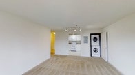 This single-floor unit presents an open-concept layout that seamlessly connects the living area to the kitchen. The living space is finished with neutral wall-to-wall carpeting and bright white walls, creating a clean, airy atmosphere. Track lighting is mounted on the ceiling, providing adjustable illumination across the main living area.

The kitchen is positioned at the far end of the main space and features white cabinetry, white appliances including an electric range and over-the-range microwave, and appears to have a tile or hard flooring surface that contrasts with the carpeted living area. The kitchen design is compact yet functional, with under-cabinet lighting adding a practical touch.

A standout feature of this unit is the in-unit stacked washer and dryer, conveniently located adjacent to the kitchen, eliminating the need for shared laundry facilities. An electrical panel is also accessible nearby. A hallway visible on the left side of the space leads to additional areas of the unit. The overall aesthetic is minimal and move-in ready, with a consistent white and neutral palette throughout.