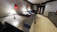 This single-floor space presents a well-appointed reception or front desk area with a contemporary design aesthetic. The focal point is a series of multi-tiered counters finished in a smooth, metallic grey surface with dark contrasting bases, arranged to accommodate multiple service points simultaneously. The countertops are accented by a digital display screen, suggesting an interactive or check-in function.

Lighting plays a key role in establishing the ambiance. Striking red cylindrical pendant lights hang from the ceiling alongside traditional white table lamps positioned along the counter, creating a layered lighting scheme that balances bold visual interest with practical illumination. Recessed ceiling lights further enhance the brightness throughout the space.

The walls feature a textured dark grey finish with crisp white trim and dark banding, lending a polished, professional appearance. The flooring consists of a patterned beige carpet that adds warmth and softens the otherwise sleek interior. In the background, glass-paneled doors allow a glimpse into adjacent areas, suggesting the space connects to further rooms or corridors. Overall, the design conveys a functional yet stylish environment suited for a hotel, office, or service-oriented facility.