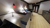 This single-floor space presents a well-appointed reception or front desk area with a contemporary design aesthetic. The focal point is a series of multi-tiered counters finished in a smooth, metallic grey surface with dark contrasting bases, arranged to accommodate multiple service points simultaneously. The countertops are accented by a digital display screen, suggesting an interactive or check-in function.

Lighting plays a key role in establishing the ambiance. Striking red cylindrical pendant lights hang from the ceiling alongside traditional white table lamps positioned along the counter, creating a layered lighting scheme that balances bold visual interest with practical illumination. Recessed ceiling lights further enhance the brightness throughout the space.

The walls feature a textured dark grey finish with crisp white trim and dark banding, lending a polished, professional appearance. The flooring consists of a patterned beige carpet that adds warmth and softens the otherwise sleek interior. In the background, glass-paneled doors allow a glimpse into adjacent areas, suggesting the space connects to further rooms or corridors. Overall, the design conveys a functional yet stylish environment suited for a hotel, office, or service-oriented facility.
