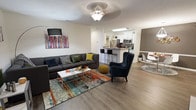 This single-floor residence offers a well-designed open-concept layout that connects the main living, kitchen, and dining areas into one cohesive and functional space. The living area is bright and inviting, featuring warm hardwood-style flooring that runs throughout the main area, adding a sense of continuity and warmth. A ceiling fan with a light fixture provides both comfort and illumination, complementing the natural light that fills the room. The living area is generously proportioned, comfortably accommodating a large sectional sofa, an accent armchair with nailhead trim, a coffee table, and a vibrant, multicolored area rug that anchors the seating arrangement. A bold piece of colorful artwork adorns the wall, adding a lively and contemporary touch to the space. The kitchen is positioned just beyond the living area, separated by a breakfast bar with seating, creating a natural transition between the two spaces. The kitchen features white cabinetry, stainless steel appliances including a refrigerator and microwave, and light countertops that give the space a clean, modern aesthetic. Adjacent to the kitchen, the dining area provides additional seating and maintains the open, airy feel of the overall layout. The combination of modern finishes, an open floor plan, and cohesive design elements gives this space a comfortable and contemporary ambiance.