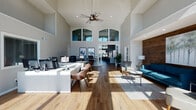 This single-floor commercial space makes an immediate impression with its dramatic double-height ceilings, creating an open and airy atmosphere throughout. Large floor-to-ceiling windows along the front facade flood the interior with natural light, offering expansive outdoor views and lending the space a bright, welcoming quality. Wide-plank hardwood flooring runs throughout, tying together the various functional zones with warmth and continuity.

The main work area features sleek, modern white desks equipped with multiple workstations, complemented by contemporary seating and ceiling fans that enhance comfort in the generously proportioned space. Arched transom windows add an architectural flourish above the interior doorways, connecting the work area to adjacent rooms with visual elegance.

A dedicated lounge zone provides a comfortable retreat, anchored by a bold teal sofa, accent chairs, and a striking horizontal shiplap wood accent wall that adds rich texture and warmth. Framed artwork and tasteful greenery contribute to an inviting, professionally curated ambiance.

Near the entrance, a pair of upholstered chairs are positioned to take full advantage of the sweeping window views, creating an ideal reception or waiting area. French doors with glass pane detailing connect the spaces, maintaining an open flow while offering privacy. The overall design balances a professional, functional layout with modern aesthetic sensibility.