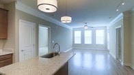 This single-floor residence offers a thoughtfully designed open-concept layout that seamlessly connects the kitchen and living areas. The kitchen is anchored by a large island with a stainless steel undermount sink and striking granite countertops featuring warm, speckled tones. Two stylish drum pendant lights hang above the island, providing focused task lighting while adding a contemporary design touch. The kitchen flows naturally into the living area, which is bathed in natural light streaming through a set of large windows dressed with horizontal blinds. A ceiling fan with a sleek fixture is centrally positioned in the living space, ensuring comfortable airflow throughout. Smooth hardwood flooring runs continuously across both areas, lending warmth and visual cohesion to the open floor plan. Crisp crown molding trims the ceilings and doorways, adding a refined architectural detail throughout. The walls are finished in a soft, muted blue-gray tone that complements the overall modern yet approachable aesthetic. A hallway visible from the kitchen leads to additional rooms, hinting at a well-organized and functional layout. Overall, the space combines practical design with clean, contemporary finishes to create a comfortable and inviting living environment.