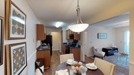 This single-floor residence offers a well-designed, open layout that seamlessly connects the kitchen, dining, and living areas. The kitchen is appointed with warm honey-toned wood cabinetry, dark countertops, and a full suite of black appliances, including a refrigerator and gas range with overhead ventilation. A stainless steel sink sits beneath cabinetry that provides generous storage throughout. The adjacent dining area is anchored by a round dark wood table with upholstered seating, illuminated by a brushed nickel pendant light with a frosted bowl shade that serves as a focal point overhead. The open sightlines between the kitchen and dining space create an inviting atmosphere well suited for both everyday meals and entertaining. Just beyond the dining area, the living room is visible, offering a comfortable and cohesive continuation of the home's neutral color palette. Warm wood-tone flooring runs throughout, tying the spaces together and adding a sense of warmth and continuity. The overall ambiance is clean, functional, and welcoming, with a practical layout that maximizes the flow between the primary living areas of the home.