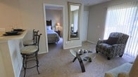 This single-floor residence offers a warm and inviting atmosphere defined by neutral wall tones and wall-to-wall carpeting that creates a cohesive, comfortable feel throughout. The living area serves as the central gathering space, furnished with a glass-topped coffee table supported by a dark wood base, a patterned upholstered chair, and an ornate wrought-iron accent chair. A large decorative mirror with a gilded frame is mounted on the wall, adding depth and visual interest to the space while reflecting ambient light from the nearby window. A vertical blind-covered window along one wall allows natural light to filter softly into the room. An open doorway connects the living area directly to the bedroom, which features plush carpeting, a substantial dark wood bed frame with a headboard, coordinating nightstands, and warm lamp lighting that contributes to the room's restful ambiance. The overall design aesthetic is traditional and understated, with carefully chosen furnishings that complement the neutral color palette and create a cohesive, livable environment throughout the home.