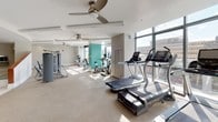 Located at 11351 Rockville Pike in Rockville, Maryland, this well-appointed fitness center offers a comprehensive workout environment designed to meet a wide range of exercise needs. The space is organized into distinct functional zones, each thoughtfully equipped and finished with a neutral color palette punctuated by vibrant teal accent walls that add a modern, energizing touch.

The main cardio area is generously proportioned, featuring a row of treadmills positioned along floor-to-ceiling windows that allow ample natural light and provide views of the surrounding urban landscape. Elliptical trainers and stationary bikes are also available, all arranged on a durable speckled flooring surface. Ceiling fans throughout the space promote air circulation, enhancing comfort during workouts.

A dedicated strength training section houses a full array of weight machines and cable equipment, while a separate free weights area includes a multi-tiered dumbbell rack, a workout bench, and large wall mirrors for form monitoring. This zone is finished with rubberized flooring suited for lifting activities.

Amenity details are not overlooked — a dedicated nook provides a modern water bottle refill station alongside wall-mounted shelving stocked with neatly rolled fresh towels, ensuring convenience for every visitor. The fitness center's curved architectural elements and open layout contribute to a clean, professional atmosphere throughout.