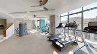 Located at 11351 Rockville Pike in Rockville, Maryland, this well-appointed fitness center offers a comprehensive workout environment designed to meet a wide range of exercise needs. The space is organized into distinct functional zones, each thoughtfully equipped and finished with a neutral color palette punctuated by vibrant teal accent walls that add a modern, energizing touch.

The main cardio area is generously proportioned, featuring a row of treadmills positioned along floor-to-ceiling windows that allow ample natural light and provide views of the surrounding urban landscape. Elliptical trainers and stationary bikes are also available, all arranged on a durable speckled flooring surface. Ceiling fans throughout the space promote air circulation, enhancing comfort during workouts.

A dedicated strength training section houses a full array of weight machines and cable equipment, while a separate free weights area includes a multi-tiered dumbbell rack, a workout bench, and large wall mirrors for form monitoring. This zone is finished with rubberized flooring suited for lifting activities.

Amenity details are not overlooked — a dedicated nook provides a modern water bottle refill station alongside wall-mounted shelving stocked with neatly rolled fresh towels, ensuring convenience for every visitor. The fitness center's curved architectural elements and open layout contribute to a clean, professional atmosphere throughout.