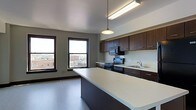 This single-floor space showcases a thoughtfully designed open-concept layout that seamlessly integrates the kitchen and living areas into one cohesive environment. The kitchen is a standout feature, outfitted with rich dark wood cabinetry that extends to the upper walls, providing generous storage throughout. A large central island with a clean, light-colored countertop serves as both a functional workspace and a natural gathering point. The kitchen is fully equipped with a stainless steel range, built-in microwave, and a sink set beneath ample counter space, all finished in matching dark cabinetry. Overhead, a combination of recessed fluorescent lighting and a pendant light ensures the space is well-illuminated. The adjacent living area benefits from multiple large windows framed in dark trim, which flood the interior with natural light and frame elevated views of the surrounding urban cityscape. The flooring is a smooth, neutral-toned surface that runs throughout, contributing to the clean and contemporary aesthetic. The overall ambiance is modern and functional, with a neutral color palette of grays and deep browns that lends the space a polished, urban character well-suited to city living.