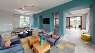 Located at 1989 Cheshire Bridge Rd NE in Atlanta's dynamic Cheshire Bridge corridor, this single-floor amenity space combines bold contemporary design with functional, welcoming zones. The lounge area makes an immediate impression with its striking teal accent walls, large-format light tile flooring, and a geometric patterned carpet in gray and yellow that anchors the seating arrangement. Plush navy swivel chairs accented with orange throw pillows surround rich wood-tone cube tables, while circular pendant lighting overhead adds a warm, modern glow. Floor-to-ceiling windows flood the space with natural light and offer open views of the surrounding greenery and streetscape. Adjacent to the lounge, a dedicated business center features a long built-in desk workspace beneath an eye-catching abstract mural in coral and teal tones. Warm wood-beamed ceiling details and pendant Edison bulbs contribute an artistic, industrial-chic character to this area, which also includes bar-height seating near additional windows. The open-plan layout connects seamlessly through teal-framed archways, allowing views across multiple functional zones including a communal gathering area with additional lounge seating and accent lighting. Throughout, the design balances energetic color with clean architectural lines, creating a polished and inviting environment.