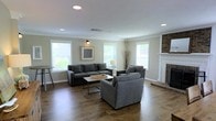 This single-floor space centers around a well-appointed living room that combines comfort with clean, contemporary design. Rich hardwood flooring extends throughout, anchoring the room with warmth and depth. Multiple windows fitted with white blinds line the walls, allowing generous amounts of natural light to brighten the neutral, soft-grey painted interior. Crown molding adds a refined architectural detail along the ceiling perimeter, complementing the recessed lighting overhead.

A classic brick fireplace with a white-painted mantle serves as a focal point on one end of the room, lending a traditional charm that balances the otherwise modern aesthetic. The living area is spacious enough to comfortably accommodate a sectional sofa and additional seating, arranged around a sleek, low-profile coffee table to create an inviting conversation area. A framed piece of wall art and a decorative floor lamp contribute subtle styling touches without overwhelming the space.

The overall ambiance is open, airy, and relaxed, making it a versatile and functional living environment suited for both everyday use and entertaining.