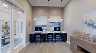 Located at 6880 Rivers Ave in North Charleston, South Carolina, this single-floor business center offers a thoughtfully designed workspace with a clean, contemporary aesthetic. The room features a built-in workstation with dark cabinetry and a light countertop, accommodating multiple desktop computers and complemented by sleek, cushioned chairs in a muted teal tone. Overhead, minimalist globe pendant lights hang from the ceiling, casting warm, focused illumination across the workspace. The walls are finished in a soft gray tone, accented by a textured wallcovering panel that serves as a refined backdrop for two large-scale abstract paintings with cool blue and beige coastal tones, adding a touch of artistic sophistication to the environment. Crown molding lines the upper perimeter of the room, lending a polished architectural detail. The flooring features a neutral, patterned carpet that ties the overall palette together seamlessly. Natural light filters in through a glass door to the left, brightening the space without disrupting its calm, professional atmosphere. The overall ambiance is modern and functional, making it well-suited for productivity and focused work in a welcoming, upscale setting.