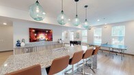 Located at 10300 University Pkwy in Pensacola, Florida, this single-floor community clubhouse is a well-appointed social hub designed for gathering, relaxing, and recreation. The open-plan layout flows seamlessly across light wood-look flooring, connecting multiple functional zones within a bright, welcoming interior.

At the heart of the space sits a substantial granite-topped island bar, illuminated by a striking row of oversized globe pendant lights in aqua-tinted glass. Warm cognac leather bar stools line the island, while a built-in counter with wood cabinetry runs along the adjacent wall, anchored by a large wall-mounted display screen. A dedicated lounge area features a row of bold yellow egg-style chairs paired with natural wood stump side tables, adding a playful, contemporary flair beneath recessed lighting.

A teal accent wall defines a secondary gathering nook with a wooden communal table and vibrant orange metal stools, illuminated by a gold multi-bulb pendant fixture. The game area includes a foosball table and a ping pong table, offering ample entertainment options. Large windows throughout the space invite abundant natural light and provide views of the surrounding outdoor landscape, including a glimpse of a pool area. Curated wall art, including a Florida-themed piece, adds local character and personality to this energetic, thoughtfully designed facility.