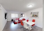 This single-story residence in Cutler Bay, Florida, presents a cohesive and boldly styled interior defined by a striking red, white, and black color palette throughout. The open-concept living and dining area serves as the heart of the home, featuring large-format gray tile flooring, a textured ceiling, and ample natural light admitted through vertical blinds. A red leather sectional sofa anchors the living space, complemented by a plush patterned area rug and a wall-mounted flat-screen television. Adjacent to the living area, a dining space accommodates a white table with red and white contemporary chairs, while a wall-mounted bar with glassware and bottle storage adds an entertaining-ready element.

The kitchen is efficiently laid out with warm wood cabinetry, granite-style countertops, a stone tile backsplash, and white appliances including a range, dishwasher, and full-size refrigerator. The bedrooms continue the monochromatic theme with black bedding, white furnishings, dark curtains, and wall-mounted televisions. Closet space is provided by louvered bi-fold doors. The bathroom features a long granite-topped vanity with dual sinks, decorative mirrors, and a drop ceiling with recessed lighting panels, offering a practical yet polished finish.