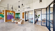 Located at 440 W State St in Columbus, Ohio, this thoughtfully designed single-floor commercial space offers a generous and open layout that balances collaborative work areas with private, enclosed rooms. Polished concrete floors flow throughout, providing a clean, modern foundation that complements the bright white ceilings with exposed structural elements and pendant lighting in a variety of styles, from sleek cylindrical fixtures to woven rattan pendants.

The open central area features a large communal wood table surrounded by rattan chairs, set atop vibrant, multicolored striped rugs that inject energy and personality into the space. Intimate lounge seating areas with light wood chairs and side tables offer additional gathering spots. Along one wall, floor-to-ceiling black-framed windows and glass doors flood the interior with natural light and provide clear sightlines to the street.

Private rooms and enclosed pods are defined by bold black steel-and-glass partition walls, maintaining visual connectivity while offering separation. Teal accent-painted nooks with bench seating and live greenery walls add a lush, biophilic touch. Warm wood-paneled feature walls display striking original artwork, including vibrant floral and botanical paintings, contributing to the space's creative and welcoming character. Tall ceilings and an abundance of natural light further enhance the sense of openness throughout.