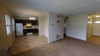 This single-floor space offers an open-concept design that seamlessly connects the living area and kitchen, creating a functional and cohesive layout. The living area is finished with wall-to-wall carpeting in a neutral beige tone, providing a comfortable and understated foundation. A floor vent provides climate control, and the walls are painted in a clean, light cream color throughout.

The kitchen is partially divided from the living space by a half-wall, maintaining an open feel while defining each area. It features dark brown cabinetry on both the upper and lower sections, offering ample storage. The countertops are light-toned, providing contrast against the darker cabinets. A white electric range and a white refrigerator are included appliances. The kitchen floor transitions to a beige tile pattern, distinguishing it from the carpeted living area. An overhead range hood is positioned above the stove, and a chandelier with multiple light fixtures hangs centrally over the kitchen, adding a decorative touch.

Toward the rear of the space, a hallway leads to additional rooms, including what appears to be a doorway to a separate room or closet. The overall ambiance is practical and move-in ready.