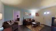 This single-floor space centers around a welcoming living area distinguished by rich, dark hardwood flooring and soft blue-painted walls that create a calm, relaxed atmosphere. The living room is furnished with a dark brown sofa accented by decorative throw pillows, paired with a matching coffee table atop a patterned area rug that adds warmth and visual interest to the space. A tall decorative vase with slender branches serves as an elegant floor accent, while wall-mounted mirrored wave décor and a colorful vintage-style painting add personality to the room. A floor lamp provides warm ambient lighting near the seating area. White trim and baseboards offer a clean contrast against the colored walls and dark flooring throughout. An open doorway leads to an adjacent room featuring distinctly lavender-pink walls and carpeted flooring, suggesting a bedroom or secondary living space. A small console table and additional furnishings are visible within that room. The entry door, positioned to the right of the living area, is framed with white molding, reinforcing the tidy, well-maintained character of this single-level space.