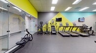 This fitness center in Ponte Vedra Beach, Florida, is a thoughtfully designed, single-floor facility offering a comprehensive range of workout options. The space is visually distinctive, featuring bold geometric wall murals in chartreuse yellow, teal, and gray that energize the environment and give the facility a modern, professional aesthetic. Durable rubber flooring throughout provides safety and comfort underfoot, while recessed panel ceiling lighting ensures consistent, bright illumination across all areas.

The main cardio zone is generously proportioned, housing multiple treadmills, elliptical trainers, stair climbers, and upright bikes, all arranged to allow comfortable spacing between equipment. Wall-mounted flat-screen televisions are positioned throughout, keeping members entertained during workouts. A dedicated strength training area features a full selection of selectorized machines targeting major muscle groups, along with benches and a cable station, set against neutral gray walls with additional mounted screens.

A separate studio room offers spin bikes, adjustable benches, and dumbbells, complemented by a large floor-to-ceiling mirror wall and a window that introduces natural light. A glass-enclosed room with bench seating, likely a sauna or steam room, adds a wellness dimension. A Matrix rowing machine and freestanding dumbbell rack round out the functional training options, making this a well-rounded facility suited for a variety of fitness goals.