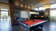 This single-floor space is designed as a contemporary recreational and social hub, combining entertainment, relaxation, and functional amenities in one cohesive layout. At the heart of the room sits a full-size pool table with a bold red felt surface, illuminated by a sleek rectangular pendant light that anchors the space visually.

The design balances dark and warm tones effectively. Dark hardwood flooring runs throughout, complemented by a dark ceiling that creates an intimate, lounge-like atmosphere. Contrasting warmly against these darker elements, a striking light wood-clad accent column serves as a bold architectural feature, adding texture and visual interest.

A comfortable lounge area with a tufted gray sectional sofa and additional seating provides a relaxed retreat adjacent to the game area. High clerestory windows along one wall introduce natural light, brightening the space without compromising its moody aesthetic.

Toward the rear, a kitchenette area features dark cabinetry, under-cabinet lighting, and what appears to be a refrigerator and sink, making the space well-suited for social gatherings. Overall, the space delivers a polished, modern ambiance ideal for recreation and entertaining.