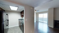 This single-floor unit offers a practical and open layout connecting the kitchen and living areas. The kitchen is outfitted with dark espresso cabinetry, dark countertops, and a stainless steel sink, complemented by tile flooring and a drop ceiling with a skylight panel that brings in additional light. A pass-through wall separates the kitchen from the main living area, creating a semi-open feel while maintaining distinct zones. The living area features dark hardwood-style flooring, a ceiling fan with light fixture, and a full-width sliding glass door that floods the space with natural light and provides direct access to an outdoor area. Neutral-toned walls throughout the unit create a clean, versatile backdrop, while textured ceilings add a classic finishing detail. The overall ambiance is functional and bright, with the contrast between the kitchen's tile flooring and the living area's wood-style flooring helping to naturally define each space. The sliding glass door serves as a focal point of the living area, offering an indoor-outdoor connection and generous natural illumination.