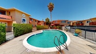 Located in the Arden-Arcade neighborhood of Sacramento, CA, this outdoor pool area serves as a central communal amenity within the residential community at 2400 Arden Way. The centerpiece is a generously sized freeform swimming pool with a distinctive kidney-like shape, finished with vivid turquoise water and classic tiled edges marked with depth indicators. The pool deck is composed of smooth concrete that extends around the perimeter, providing ample space for lounging and movement. A dedicated seating area is furnished with cushioned red chairs, a loveseat, a coffee table, and shaded by large red market umbrellas, creating a comfortable outdoor living environment. The area is enclosed by a wrought iron fence and neatly trimmed hedgerows that offer both security and a sense of privacy. Lush tropical landscaping, including a towering palm tree and various shrubs, adds natural character to the setting. The surrounding two-story residential buildings feature warm stucco facades in earthy orange tones with red accents and balconies that overlook the pool area. Under the abundant California sunshine, this outdoor space presents a bright, vibrant, and inviting atmosphere ideal for relaxation and recreation.