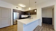 Located at 3100 Park Ave in Tustin, CA, this one-bedroom, one-bathroom residence offers a thoughtfully designed layout that maximizes every square foot. The open-concept kitchen and living area serves as the heart of the home, featuring a large peninsula with a crisp white countertop, dark espresso cabinetry, stainless steel appliances including a refrigerator, microwave, and gas range, and stylish pendant lighting overhead. Wood-style plank flooring runs throughout the main living areas, complementing the modern, neutral color palette. The living room is bright and airy, benefiting from natural light through a sliding glass door that opens to a private balcony. A hallway connects the living space to the private quarters, where the bedroom offers plush carpeting, a window with blinds for natural light and privacy, and direct access to a walk-in closet complete with built-in shelving and hanging rods. The full bathroom is clean and functional, featuring wood-style flooring, a tiled shower/tub combo, built-in open shelving, and a dark-cabineted vanity with a white countertop. A dedicated hallway provides convenient circulation throughout the unit, rounding out this well-appointed Tustin residence.