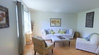 This two-story space offers a clean, airy aesthetic defined by light hardwood flooring and soft, neutral wall tones that create a calm and inviting atmosphere throughout. The living area is a standout feature, showcasing a generously sized white sectional sofa dressed with an assortment of blue, green, and patterned throw pillows, complemented by a tufted rectangular ottoman that doubles as a central coffee table. A patterned accent chair in muted gold and grey adds a touch of visual interest to the seating arrangement. Natural light filters in through a large window dressed with sheer white curtains, brightening the space and enhancing its open, relaxed feel. A framed floral artwork in warm orange and yellow tones serves as a focal point on the main wall, adding a subtle pop of color against the pale backdrop. Decorative branches and a small side console near the window contribute understated organic touches to the room. The overall ambiance is light, casual, and well-curated, balancing comfort with a cohesive, welcoming design sensibility across both floors.
