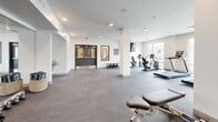 This fitness center in Phoenix, Arizona offers a spacious, single-floor exercise room designed to support a comprehensive workout experience. The layout is open and thoughtfully organized, providing ample room to move between stations without feeling crowded.

The cardio area is positioned near a wall of large windows that flood the space with natural light and offer views of the surrounding exterior, creating an energizing atmosphere for treadmill and elliptical workouts. Multiple treadmills and an elliptical trainer are available in this bright zone.

The strength and conditioning area features a diverse selection of equipment, including weight machines, a rowing machine, a stationary bike, a flat bench, dumbbells, and kettlebells. A full-length mirror spans one wall, enhancing the sense of depth and allowing users to monitor their form during exercise.

A standout design element is the striking desert-inspired mural adorning the back wall — a sweeping landscape of warm golden hills beneath a soft sun in muted tones of terracotta, sand, and sky blue. This artistic feature adds a distinctive and welcoming character to the space.

The flooring throughout is durable rubber-style gym flooring in a neutral gray, complementing the clean white walls and recessed lighting overhead. A water fountain near the entrance adds a practical convenience. The overall ambiance is modern, clean, and motivating.