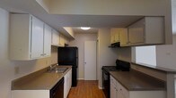 This single-story space centers around a well-organized galley-style kitchen designed to maximize functionality within a compact layout. The kitchen features a parallel configuration, with countertops and cabinetry lining both sides of a central corridor, creating an efficient workflow for cooking and meal preparation. White upper and lower cabinets provide ample storage, complemented by dark laminate countertops that offer a practical contrast. Black appliances, including a refrigerator, electric range, and dishwasher, are integrated seamlessly into the design. A stainless steel single-basin sink is positioned along one wall, accompanied by under-cabinet space below. Overhead, flush-mount lighting provides consistent illumination throughout the kitchen. The flooring consists of warm-toned hardwood, adding a grounded, natural aesthetic to the space. At the far end of the kitchen, a white door leads to an adjoining room or utility area, suggesting additional functional space beyond. The overall ambiance is clean, straightforward, and practical, suited for everyday use.