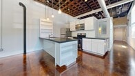 Located at 3502 Scotts Lane in Philadelphia, PA, this loft-style unit offers a striking combination of historic architectural details and contemporary design. The single-floor layout features rich, acid-stained polished concrete floors throughout, which catch and reflect light beautifully, adding warmth and depth to every room.

The open-concept kitchen and living area is a standout space. The kitchen is equipped with stainless steel appliances, including a refrigerator, range, and built-in microwave, all complemented by crisp white cabinetry, dark stone countertops, and a central island with a sink and open shelving beneath. Pendant lighting hangs from an exposed coffered ceiling structure, which, along with visible ductwork, reinforces the industrial-loft aesthetic.

The living area and an additional room showcase the building's storied past through patches of original stone masonry walls that have been intentionally left exposed, framed by white-painted plaster. Natural light enters through multiple windows fitted with blinds, illuminating the textured surfaces throughout. The open ceiling grid and industrial ductwork add to the raw, loft-like character of the space.

The bathroom features a generously sized soaking tub surrounded by neutral tile surround, offering a clean and calming retreat. Overall, this unit masterfully balances historic charm with modern livability.