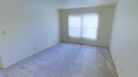 This single-floor space presents a clean, understated interior designed for comfortable everyday living. The main room is generously proportioned, featuring wall-to-wall plush carpeting in a light neutral tone that adds warmth and softness underfoot. The walls are finished in a crisp off-white, contributing to a bright and airy atmosphere throughout the space.

A large window dressed with vertical blinds serves as a focal point of the room, allowing an abundance of natural light to filter in while offering adjustable privacy control. The blinds diffuse the incoming light evenly, casting a soft, even glow across the room. A floor vent is visible near the baseboard, indicating a centralized climate control system for year-round comfort.

The overall layout is open and flexible, providing ample room for a variety of furniture arrangements to suit different lifestyle needs. The neutral color palette and clean lines of the space create a versatile backdrop that can easily accommodate a range of interior design styles. The single-floor layout ensures convenient accessibility throughout.