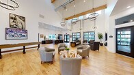 Located at 6705 Linderson Way SW in Tumwater, Washington, this single-floor community clubhouse offers a generously proportioned, open-plan interior designed for both social gathering and relaxed lounging. The space is immediately distinguished by its dramatically soaring ceilings, accented by exposed wooden beams and industrial-style ductwork that lend a modern yet warm character throughout.

Natural light pours in through expansive floor-to-ceiling windows and large glass doors, illuminating the wide-plank hardwood floors that extend across the entire space. A striking collection of statement light fixtures — including oversized ring chandeliers and starburst pendants — adds a sophisticated, curated aesthetic.

The main lounge area features multiple seating arrangements, including plush tufted sofas, upholstered barrel chairs, and a distinctive live-edge wood coffee table, all centered around a wall-mounted television with a linear fireplace below. A dedicated game area with a foosball table and shuffleboard provides additional recreational amenities.

The fully equipped kitchen showcases dark wood cabinetry, white quartz countertops, a large center island with bar seating, stainless steel appliances, and a classic white subway tile backsplash illuminated by industrial pendant lighting. The welcoming entrance foyer features tall windows, statement lighting, and direct leasing office access.
