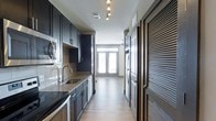 This thoughtfully designed single-floor space offers a seamless blend of modern style and functional living. The galley-style kitchen is a standout feature, outfitted with deep navy cabinetry, granite countertops, and a full suite of stainless steel appliances, including a built-in microwave, dishwasher, and refrigerator. A subway tile backsplash adds a clean, contemporary touch, while track lighting overhead ensures the workspace is well-illuminated. The undermount sink with a brushed nickel faucet adds a refined detail to the overall design. Light hardwood-style flooring runs throughout, creating a warm and cohesive aesthetic that connects the kitchen to the living areas beyond. At the end of the corridor, a set of French doors floods the space with natural light, opening the interior to an abundance of brightness and providing a view beyond. A ceiling fan is visible in the adjoining living area, enhancing comfort and airflow. A louvered door along the corridor provides additional storage access, contributing to the space's practical layout. The overall ambiance is modern and polished, with dark contrasting tones balanced by light flooring and generous natural light.