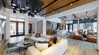 This single-floor space showcases a thoughtfully designed modern interior that seamlessly blends communal lounging, socializing, and productivity. The open-concept layout is anchored by wide-plank hardwood flooring and a striking ceiling treatment that combines warm wood panels with exposed beams and bold black structural accents, creating a layered visual depth throughout.

The main lounge area is generously proportioned, featuring oversized cream sectional sofas accented with rich jewel-toned throw pillows, arranged around a wall-mounted display screen set against a dark stone feature wall. Dramatic cluster pendant lighting in matte black and brass finishes serves as a defining design element overhead. Large black-framed floor-to-ceiling windows flood the space with natural light while offering views of the surrounding streetscape.

A dedicated study and workspace nook is finished with warm wood built-in shelving, a black countertop desk, textured carpet flooring, and leather lounge chairs. An artistic watercolor mural adds a creative focal point to this zone. Additional intimate seating areas with cognac leather chairs are positioned near oversized windows, ideal for reading or quiet conversation.

French doors lead to an exterior entry, and the overall ambiance balances industrial-modern architecture with warm, residential-inspired furnishings throughout.