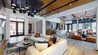 This single-floor space showcases a thoughtfully designed modern interior that seamlessly blends communal lounging, socializing, and productivity. The open-concept layout is anchored by wide-plank hardwood flooring and a striking ceiling treatment that combines warm wood panels with exposed beams and bold black structural accents, creating a layered visual depth throughout.

The main lounge area is generously proportioned, featuring oversized cream sectional sofas accented with rich jewel-toned throw pillows, arranged around a wall-mounted display screen set against a dark stone feature wall. Dramatic cluster pendant lighting in matte black and brass finishes serves as a defining design element overhead. Large black-framed floor-to-ceiling windows flood the space with natural light while offering views of the surrounding streetscape.

A dedicated study and workspace nook is finished with warm wood built-in shelving, a black countertop desk, textured carpet flooring, and leather lounge chairs. An artistic watercolor mural adds a creative focal point to this zone. Additional intimate seating areas with cognac leather chairs are positioned near oversized windows, ideal for reading or quiet conversation.

French doors lead to an exterior entry, and the overall ambiance balances industrial-modern architecture with warm, residential-inspired furnishings throughout.