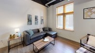 This single-floor space combines industrial design elements with modern finishes to create a functional and stylish living environment. The living area is a standout feature, boasting soaring ceilings with exposed black structural beams and ductwork, warm wood-look flooring, and large windows with wood-framed blinds that invite abundant natural light. The space accommodates both a sectional seating area and a separate lounge corner with a ceiling fan overhead.

The kitchen is well-appointed with white shaker-style cabinetry extending to the ceiling, a classic white subway tile backsplash, and a full suite of stainless steel appliances including a large French door refrigerator, electric range with microwave, and ample counter space.

Multiple bedrooms offer comfortable accommodations, each with wood-look flooring and natural light from windows with wood-framed blinds. One bedroom includes a dedicated closet with built-in shelving and hanging rods for added storage.

Two bathrooms feature clean white walls, wood-look flooring, and sleek matte black fixtures and hardware for a contemporary touch. The dedicated laundry room houses a full-size top-load washer and dryer pair beneath generous white upper cabinetry, providing convenient in-unit laundry and additional storage.