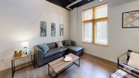 This single-floor space combines industrial design elements with modern finishes to create a functional and stylish living environment. The living area is a standout feature, boasting soaring ceilings with exposed black structural beams and ductwork, warm wood-look flooring, and large windows with wood-framed blinds that invite abundant natural light. The space accommodates both a sectional seating area and a separate lounge corner with a ceiling fan overhead.

The kitchen is well-appointed with white shaker-style cabinetry extending to the ceiling, a classic white subway tile backsplash, and a full suite of stainless steel appliances including a large French door refrigerator, electric range with microwave, and ample counter space.

Multiple bedrooms offer comfortable accommodations, each with wood-look flooring and natural light from windows with wood-framed blinds. One bedroom includes a dedicated closet with built-in shelving and hanging rods for added storage.

Two bathrooms feature clean white walls, wood-look flooring, and sleek matte black fixtures and hardware for a contemporary touch. The dedicated laundry room houses a full-size top-load washer and dryer pair beneath generous white upper cabinetry, providing convenient in-unit laundry and additional storage.