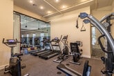 This single-level fitness center is thoughtfully designed to accommodate a comprehensive range of workout routines. The space is divided into two interconnected zones, seamlessly accessible to one another. The cardio area is anchored by multiple treadmills and an elliptical trainer, all positioned along a full-height glass wall partition that offers a clear view into a furnished lounge area beyond, creating an open, airy atmosphere. Recessed lighting and exposed ceiling tracks illuminate the space evenly throughout.

The strength and functional training zone features a multi-station cable machine, a stationary cycle, a recumbent bike, and an adjustable bench, providing ample options for resistance and conditioning work. Neutral-toned carpet covers the floors throughout both areas, and the crisp white walls enhance the sense of brightness. A window in the strength zone allows natural light to filter in, offering views of surrounding greenery.

A decorative patterned tile near the entryway adds a subtle design accent, while wall-mounted resistance equipment provides additional training versatility. The overall layout is clean, functional, and well-maintained, making it a practical and inviting environment for daily fitness use.