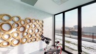 This contemporary fitness studio is thoughtfully designed to offer an inspiring and functional workout environment. The space is anchored by a set of stationary cycling bikes, positioned to take full advantage of the room's most striking architectural feature: expansive floor-to-ceiling windows fitted with sleek black frames. These large glass panels flood the interior with abundant natural light and open up to sweeping views of the surrounding urban landscape, including a terrace or balcony area beyond the glass doors.

One wall is adorned with a bold, sculptural installation composed of interlocking circular wooden forms in a warm golden tone, adding an artistic focal point that balances the room's clean, neutral palette. The walls are finished in a soft, textured white, complementing the light grey carpeted flooring that runs throughout the space.

A full-length mirror along one wall visually expands the room and reflects both the equipment and the natural light, enhancing the sense of openness. Recessed and track lighting in the coffered ceiling provides ample illumination for any time of day. A wall-mounted speaker and audio controls support a motivating workout atmosphere. The natural wood entry door adds a warm, refined touch to the overall modern aesthetic.