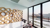 This contemporary fitness studio is thoughtfully designed to offer an inspiring and functional workout environment. The space is anchored by a set of stationary cycling bikes, positioned to take full advantage of the room's most striking architectural feature: expansive floor-to-ceiling windows fitted with sleek black frames. These large glass panels flood the interior with abundant natural light and open up to sweeping views of the surrounding urban landscape, including a terrace or balcony area beyond the glass doors.

One wall is adorned with a bold, sculptural installation composed of interlocking circular wooden forms in a warm golden tone, adding an artistic focal point that balances the room's clean, neutral palette. The walls are finished in a soft, textured white, complementing the light grey carpeted flooring that runs throughout the space.

A full-length mirror along one wall visually expands the room and reflects both the equipment and the natural light, enhancing the sense of openness. Recessed and track lighting in the coffered ceiling provides ample illumination for any time of day. A wall-mounted speaker and audio controls support a motivating workout atmosphere. The natural wood entry door adds a warm, refined touch to the overall modern aesthetic.