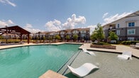 Silver Oaks offers a thoughtfully designed outdoor amenity space centered around a large resort-style swimming pool with crystal-clear turquoise water. The pool area is surrounded by a generously sized sundeck outfitted with lounge chairs, providing ample space for sunbathing and relaxation beneath open skies.

Adjacent to the pool is a covered outdoor pavilion that serves as the social heart of the community. The pavilion features soaring vaulted ceilings with exposed dark wood beam accents, white shiplap paneling, and ceiling fans to keep the space comfortable year-round. Large sets of glass French doors connect the pavilion to an interior clubhouse, allowing seamless indoor-outdoor flow and views of the pool beyond.

The covered lounge area is furnished with plush seating arrangements — including sofas and accent chairs — centered around a flat-screen television mounted overhead, making it an ideal gathering spot. String lights draped from the pergola structure add a warm, inviting ambiance during evening hours.

A fully equipped outdoor kitchen is integrated into the pavilion, featuring a built-in grill, stainless steel countertops, a beverage cooler, and pendant lighting, all set against a classic brick surround. Manicured landscaping with trimmed hedges and mature trees frames the entire outdoor space, creating a lush, resort-like atmosphere throughout.