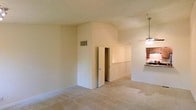 This two-floor residence offers a welcoming and functional layout designed for comfortable everyday living. The main living area is characterized by vaulted, arched ceilings that lend a sense of height and openness to the space. Soft, neutral-toned walls are complemented by plush wall-to-wall carpeting, creating a warm and inviting atmosphere throughout.

A key feature of the layout is the open sightline connecting the living area to the kitchen, which is partially visible and separated by a raised bar-height counter — ideal for casual dining or entertaining. The kitchen is fitted with dark wood cabinetry and benefits from a pendant light fixture that adds a touch of warmth. A ceiling fan in the transitional space between the living and kitchen areas provides both functionality and additional ambient lighting.

The living area also includes a door leading to an adjoining room or hallway, offering well-considered separation of spaces while maintaining the open feel of the overall floor plan. A ventilation grille positioned high on the interior wall reflects a thoughtfully designed HVAC system integrated into the architecture. The two-floor configuration provides additional living space, making this a versatile and practical residence suited to a variety of lifestyles.