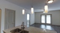 This single-floor residence showcases a thoughtfully designed open-concept layout that seamlessly connects the kitchen and living areas. The kitchen is anchored by a sleek granite countertop island with a double stainless steel sink and a gooseneck faucet, providing both functionality and style. Overhead, a trio of cylindrical pendant lights hang from the elevated ceiling, casting a warm, ambient glow across the space while reinforcing the modern aesthetic.

The living area extends naturally from the kitchen, featuring rich hardwood-style flooring that runs throughout. A ceiling fan with an integrated light fixture provides both comfort and additional illumination. Natural light flows generously into the space through multiple windows fitted with horizontal blinds, as well as a glass-paneled door that leads to an exterior access point, further brightening the interior.

The neutral gray walls and clean architectural lines create a contemporary yet inviting atmosphere. A secondary room visible through an interior doorway benefits from its own window, offering additional natural light. The overall design balances practicality with modern finishes, making this a well-appointed and functional living space with a cohesive, understated style throughout.