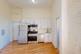 Located at 125 Magnolia Avenue in Jersey City, NJ, this efficiently designed unit makes a strong architectural impression despite its compact footprint. The main living area is defined by soaring ceilings that amplify the sense of space, warm hardwood flooring throughout, and a striking whitewashed exposed brick wall that adds texture and character. A large window flanked by the brick wall allows natural light to pour in, illuminating the room and highlighting a classic cast-iron radiator beneath it — a nod to the building's historic charm.

The kitchen is thoughtfully laid out along one wall, featuring white cabinetry that extends nearly to the ceiling, stainless steel appliances including a gas range, built-in microwave, refrigerator, and dishwasher, as well as a clean white countertop with an integrated sink. The exposed brick detail continues into the kitchen area, providing a cohesive aesthetic.

The bathroom is clean and modern, finished with white subway tile walls, hexagonal mosaic floor tile, a walk-in shower, a compact vanity with an oval mirror, and a toilet — all presented in a crisp, all-white palette. Every element of this unit reflects a deliberate blend of historic industrial character and contemporary finishes.